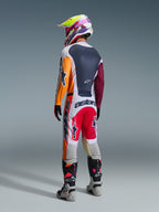 Motocross-Fahrer in kompletter Rennausrüstung von hinten, Alpinestars 2026 Racer Portl Trikot in Rot, Hellgrau und Dunkelgrün mit orangefarbenen und kastanienbraunen Einsätzen, passende weiß-rote Hose, grau-schwarze Stiefel mit rosa Details, mehrfarbiger Helm, abgestimmtes Offroad-Rennoutfit