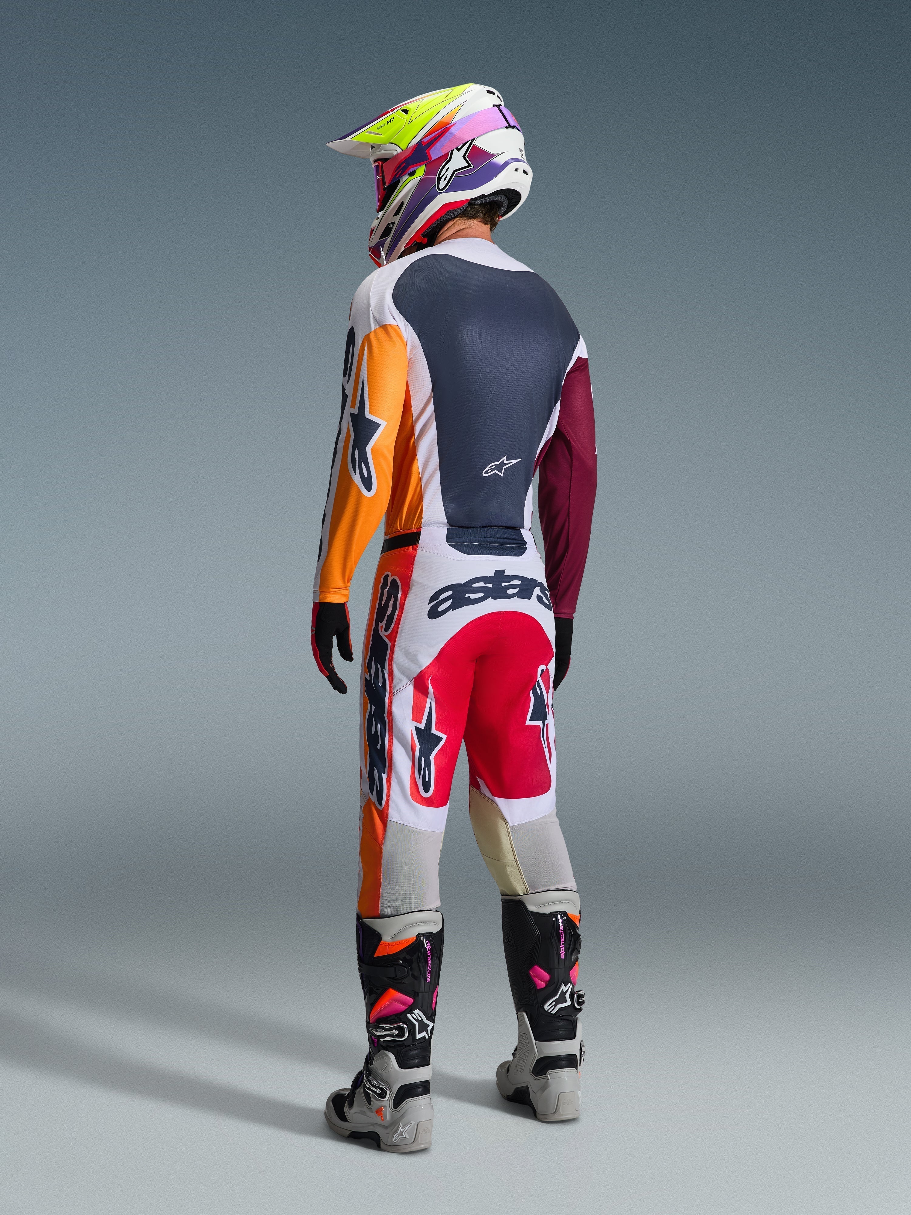 Motocross-Fahrer in kompletter Rennausrüstung von hinten, Alpinestars 2026 Racer Portl Trikot in Rot, Hellgrau und Dunkelgrün mit orangefarbenen und kastanienbraunen Einsätzen, passende weiß-rote Hose, grau-schwarze Stiefel mit rosa Details, mehrfarbiger Helm, abgestimmtes Offroad-Rennoutfit