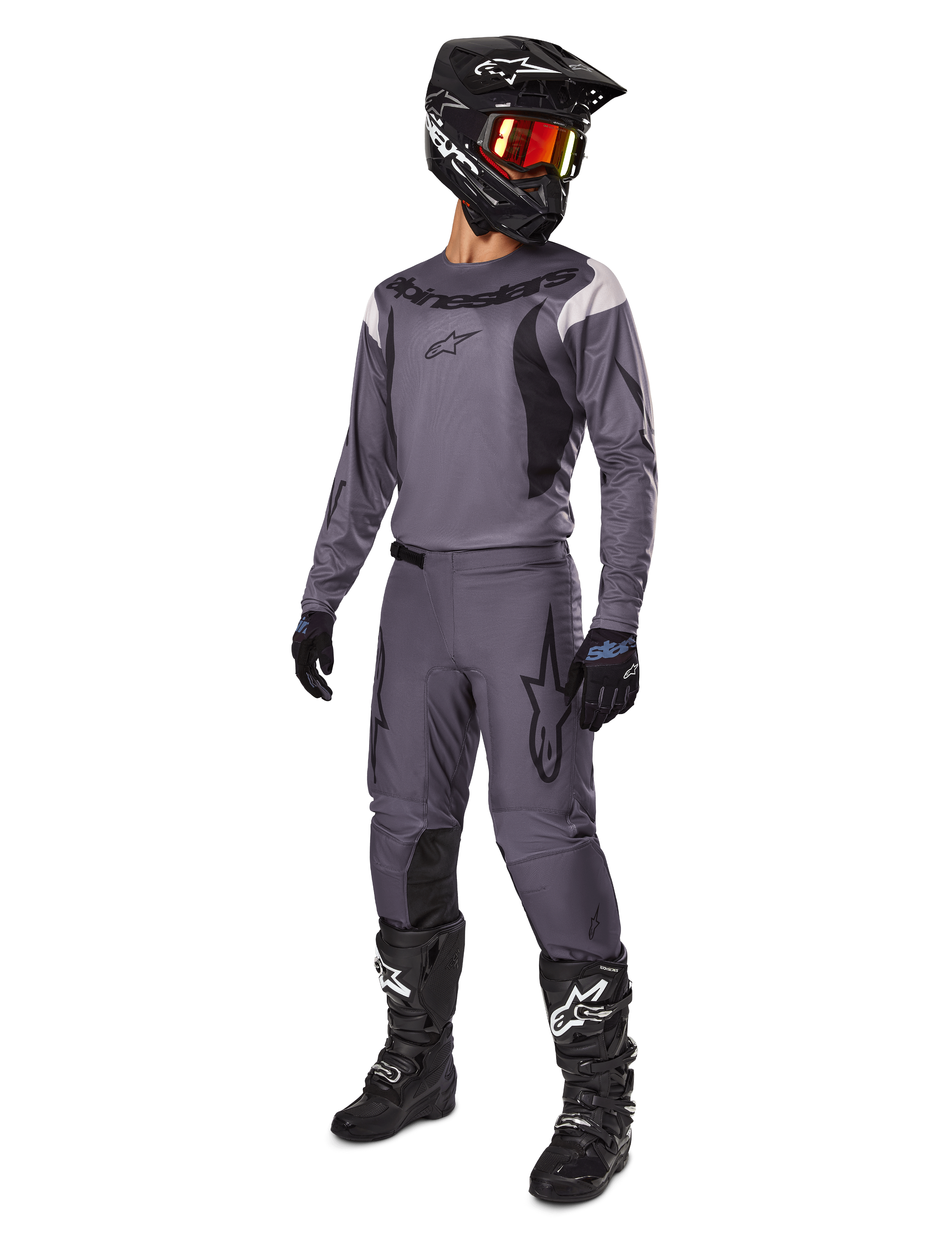 Motocross-Fahrer im Alpinestars 2025 Fluid Haul Ausrüstungsset in Grau, inklusive grauem Jersey und Hose mit schwarzen Akzenten, schwarzem Helm mit rötlicher Brille, schwarzen Handschuhen und schwarzen Stiefeln mit weißen Logos, komplettes Offroad-Rennoutfit
