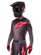 Alpinestars 2025 Supertech Lipan Gear Set, Motocross rider wearing a dark grey jersey and pants with red and white accents, featuring a large red Alpinestars logo on the chest, laser-cut perforations for ventilation, matching black helmet, red goggles, and red and black gloves.