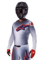Motocross-Fahrer im Alpinestars 2025 Supertech Lipan Ausrüstungsset, graues Trikot und Hose mit navyblauen und orangefarbenen Akzenten, mattschwarzer Helm mit Brille, rote und schwarze Handschuhe, sportliche Performance-Ausrüstung für Offroad-Rennsport