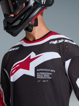 Alpinestars 2026 Supertech AFD Vista Trikot, Nahaufnahme eines Motocross-Fahrers in einem schwarzen Jersey mit roten und weißen Akzenten, mit einem großen weiß-roten Alpinestars-Logo, lasergeschnittenen Belüftungsperforationen und professioneller Rennpassform, kombiniert mit einem schwarzen Helm.