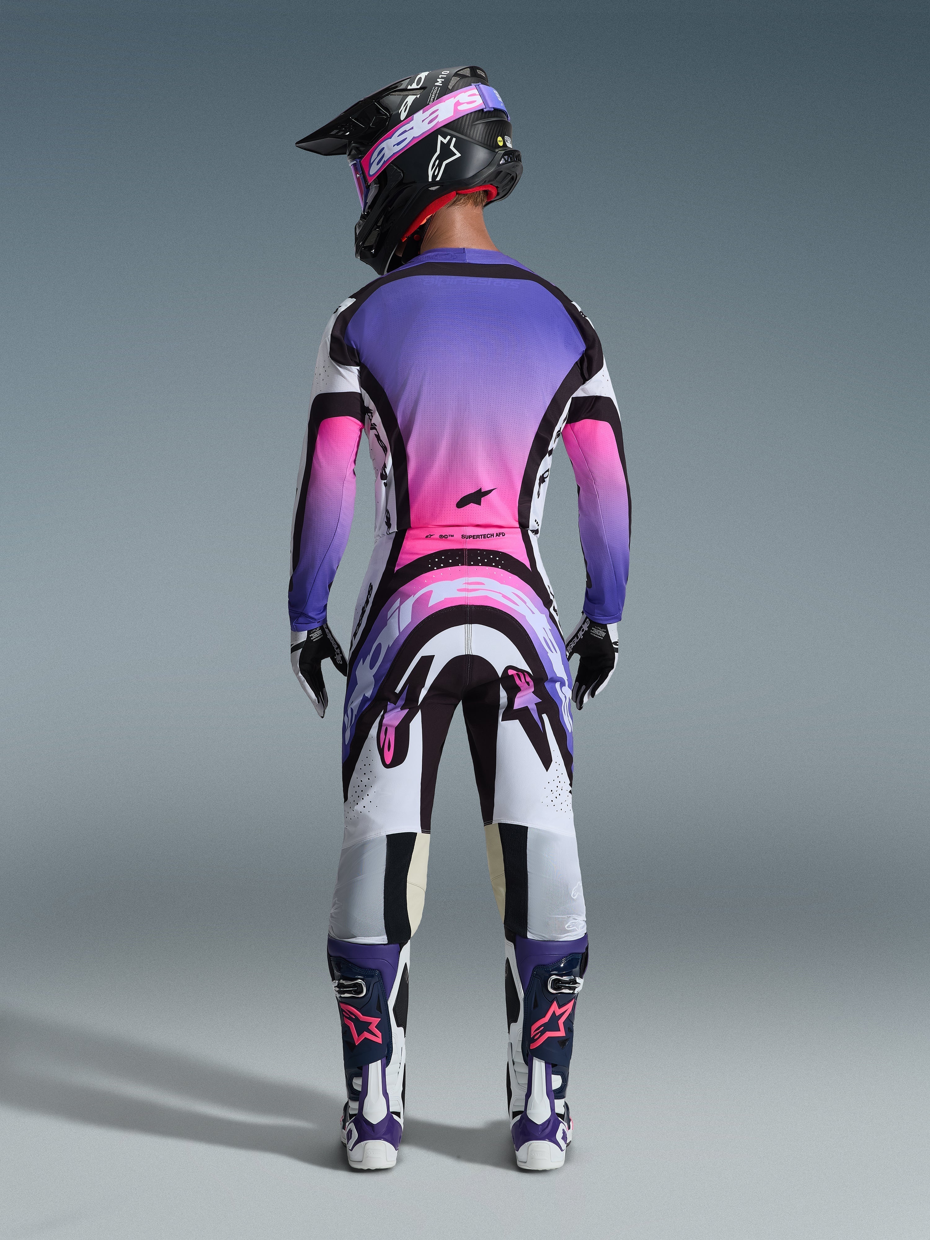 Motocross-Fahrer in der Rückansicht mit dem 2026 Supertech AFD Vista Trikot, Weiß, Lila und Schwarz mit pinkfarbenen Farbverlauf-Akzenten, passende Motocross-Hose, schwarzer Helm mit pinker Brille und weiß-lila Stiefel, komplettes professionelles Rennausrüstungs-Set