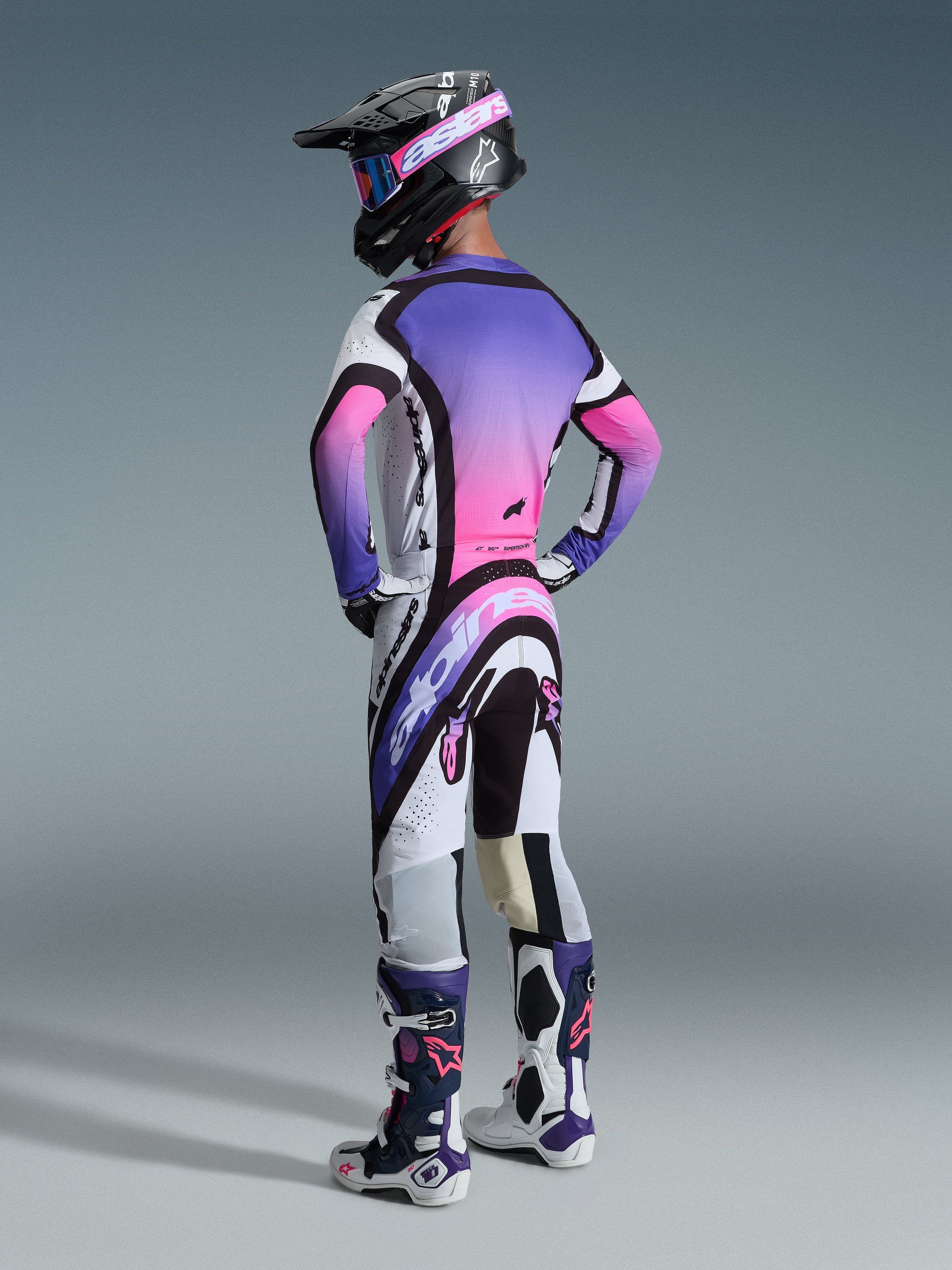 Alpinestars 2026 Supertech AFD Vista Trikot, Motocross-Trikot, Weiß, Lila und Schwarz mit pinkfarbenem Farbverlauf, von hinten getragen mit passender Motocross-Hose, schwarzem Helm und weiß-lila Stiefeln, zeigt die komplette professionelle Rennausrüstung für Offroad-Performance.
