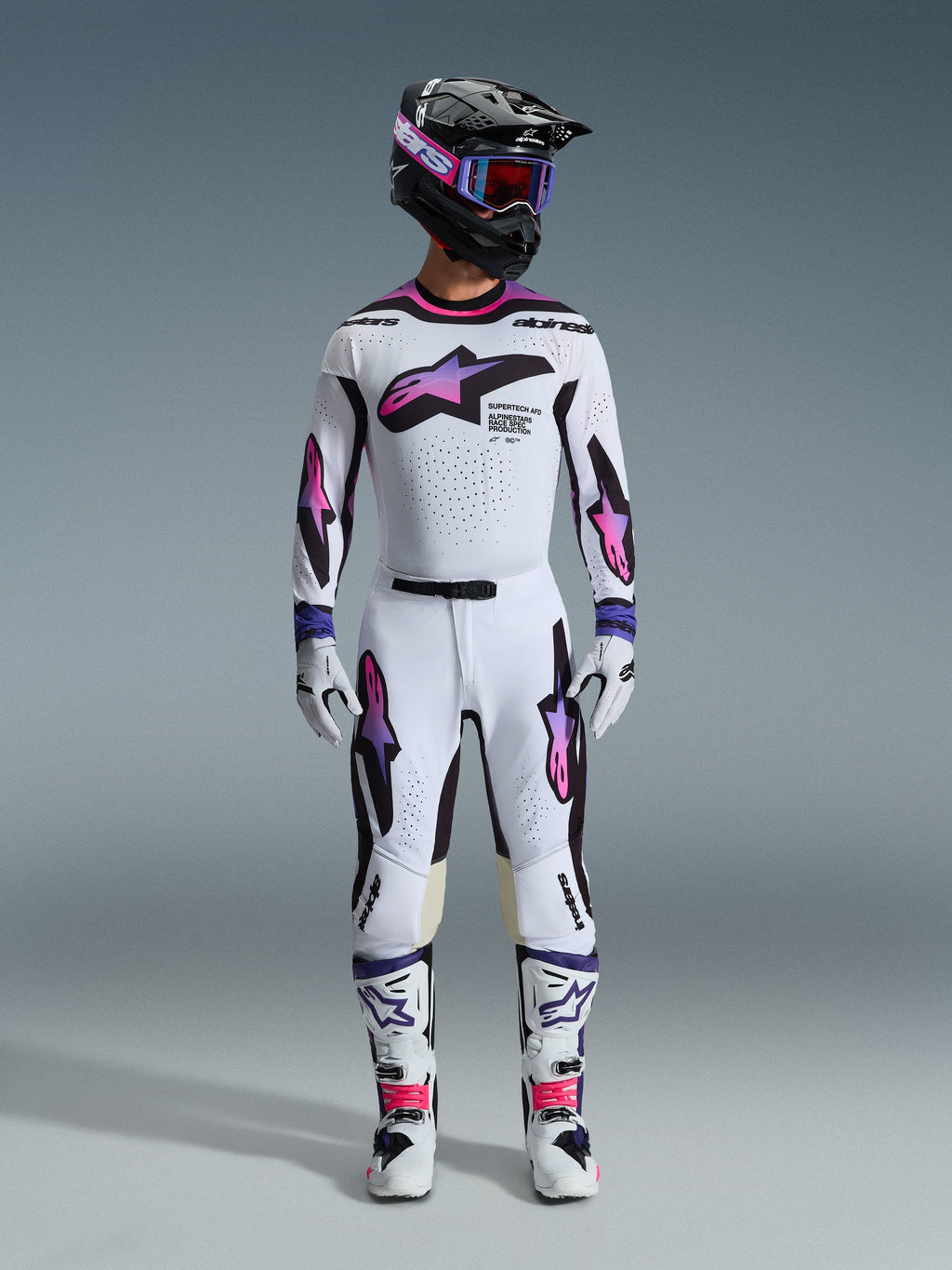 Motocross-Fahrer in kompletter Rennausrüstung, Alpinestars 2026 Supertech AFD Vista Trikot in Weiß Lila Schwarz, passende weiße Hose mit lila-rosa Farbverlauf-Grafiken, weiße Stiefel, schwarzer Helm mit lila Brille, professionelles Offroad-Rennoutfit