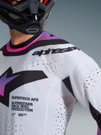 Alpinestars 2026 Supertech AFD Vista Trikot, Motocross-Trikot, Weiß, Lila und Schwarz, Detailansicht mit lasergeschnittenen Perforationen und Race-Spec-Branding, getragen mit einem schwarzen Helm, professionelle Offroad-Rennausrüstung