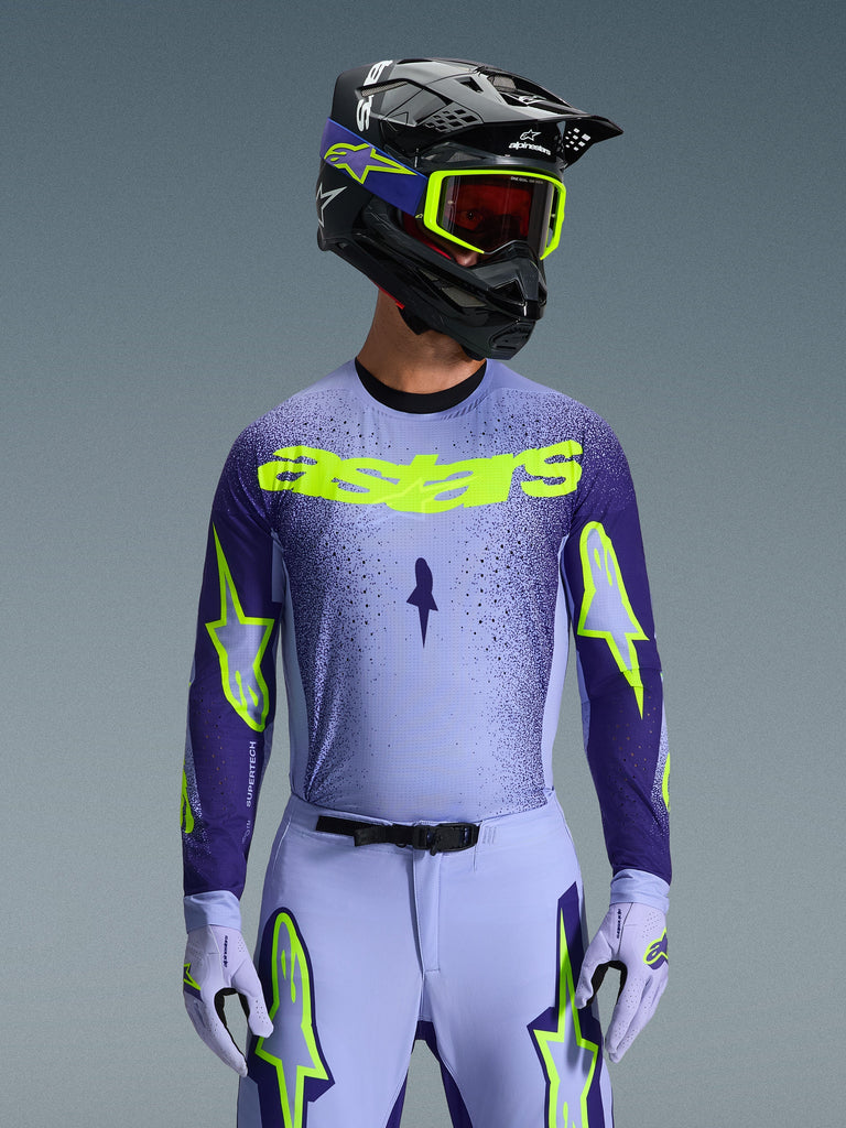 Alpinestars 2026 Supertech Scenz Jersey, Motocross-Trikot, Lila und Neongelb, getragen von einem Fahrer mit passendem schwarz-lila Helm, neongelber Brille und lila Rennhose, mit neongelbem Astars-Branding auf der Brust und Stern-Logos auf den Ärmeln.