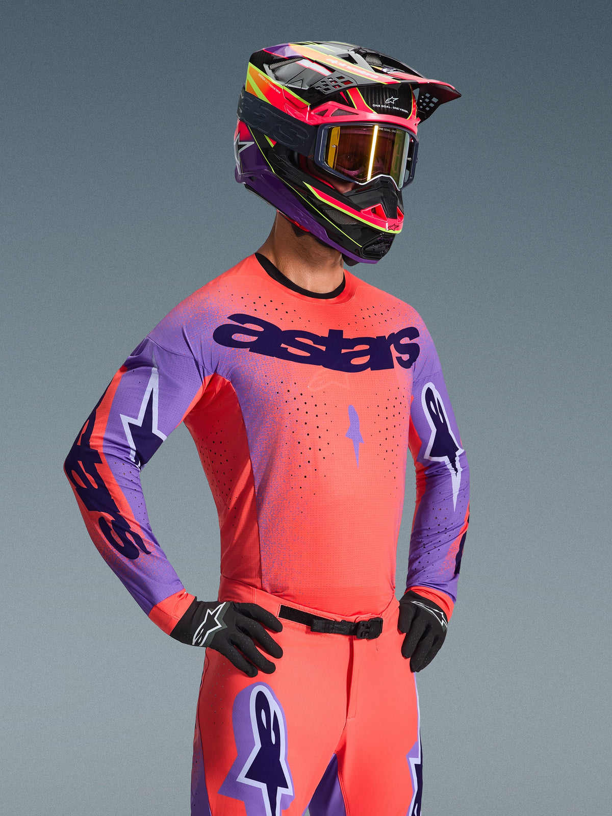 Motocross-Fahrer im Alpinestars 2026 Supertech Scenz Jersey, Hot Coral und Lila mit markantem 'astars'-Branding auf der Brust, passender Ausrüstung inklusive korallenfarbener Hose und einem abgestimmten lila-korallenfarbenen Helm, mit lasergeschnittenen Perforationen für Hochleistungs-Offroad-Rennsport.