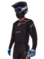 Motocross-Fahrer im Alpinestars 2025 Racer Graphite Ausrüstungsset, schwarzes Jersey mit grauen Alpinestars-Logos, passende schwarze Hose mit grauen Akzenten, schwarzer Helm mit blau-weißer Brille, blau-graue Handschuhe, elegantes technisches Outfit für Offroad-Rennsport