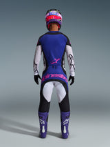 Rear view of a motocross rider wearing the Alpinestars 2026 Techstar Knif Jersey in Black, Light Gray, and Purple, featuring white sleeves and a pink logo on the lower back. Paired with matching motocross pants, purple boots, and a colorful helmet in a studio setting.
