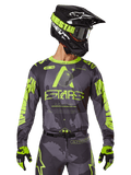 Alpinestars 2025 Racer Hollow Trikot, Motocross-Renntrikot, Dunkles Camo und Neongelb, getragen mit passender Hose, schwarz-gelbem Helm mit Brille und schwarzen Handschuhen, mit Alpinestars-Branding und Camouflage-Design