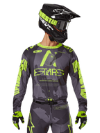Alpinestars 2025 Racer Hollow Trikot, Motocross-Renntrikot, Dunkles Camo und Neongelb, getragen mit passender Hose, schwarz-gelbem Helm mit Brille und schwarzen Handschuhen, mit Alpinestars-Branding und Camouflage-Design