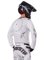 Motocross-Fahrer in der Rückansicht trägt das Alpinestars 2025 Racer Hollow Gear Set in Weiß, weißes Langarmtrikot mit grauen Sternlogos und kreisförmigen Grafiken, passende weiße Hose mit markantem Branding am Gesäß, schwarzer Helm, abgestimmte Offroad-Rennausrüstung.