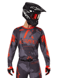 Alpinestars 2025 Racer Hollow Jersey, Motocross-Trikot, Gray Camo und Orange Fluo, getragen mit passendem Ausrüstungsset inklusive Hose und Handschuhen, schwarzer Helm mit weißen Grafiken und Schutzbrille, markantes ASTARS-Branding, schützende Offroad-Rennsportbekleidung