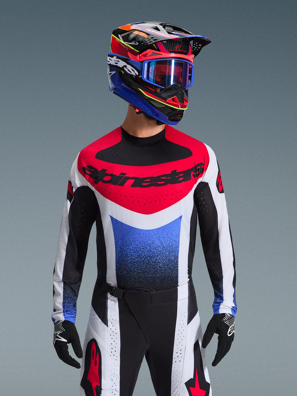 Alpinestars 2026 Techstar Knif Gear Set, motocross rider wearing complete racing gear, red white and blue jersey with large black branding, matching black and white pants with red star logo, multi-colored helmet with blue goggles and black gloves, coordinated outfit for off-road competition