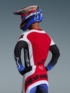 Motocross rider viewed from behind wearing the 2026 Techstar Knif Jersey in Red White and Black, featuring laser-cut perforations on the back and bold branding, paired with matching pants with blue accents and a blue and black Alpinestars helmet, professional off-road racing aesthetic