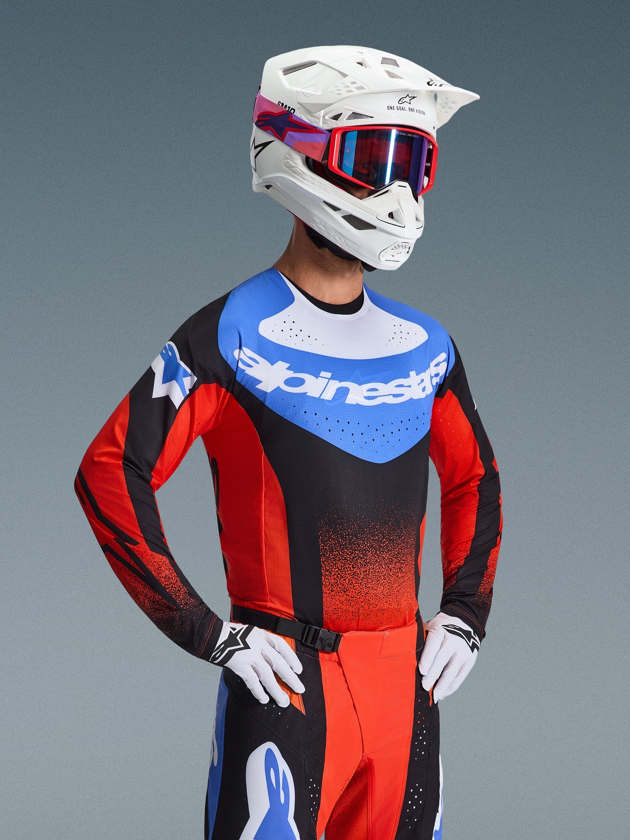 Motocross-Fahrer trägt das 2026 Techstar Knif Jersey in Orange, Schwarz und UCLA-Blau, mit einem blauen Brusteinsatz mit weißem Alpinestars-Logo, schwarzen und orangefarbenen Ärmeln mit lasergeschnittenen Perforationen, kombiniert mit einem weißen Helm, einer roten Goggle und passender Rennhose.
