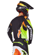 Motocross-Fahrer von hinten im Alpinestars 2025 Fluid Wurx Ausrüstungsset, schwarzes Trikot mit leuchtenden neongelben und orangefarbenen Grafiken, passende Hose mit weißen und orangefarbenen Einsätzen, schwarzer Helm mit Neon-Akzenten, markantes Alpinestars-Branding auf den Ärmeln und am Gesäß.