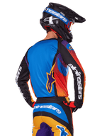 Motocross-Fahrer in der Rückansicht, Alpinestars 2025 Fluid Wurx Ausrüstungsset, mehrfarbiges Trikot mit blauem Rücken und schwarzen Ärmeln mit weißen Logos, passende orange-blaue Hose, schwarz-blauer Helm mit orangefarbener Schutzbrille, koordiniertes Offroad-Rennoutfit.