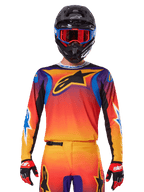 Alpinestars 2025 Fluid Wurx Jersey, mehrfarbiges Motocross-Trikot mit orange-rotem Farbverlauf und lila Akzenten, getragen mit passender Ausrüstung, schwarzer Helm mit blau-roter Brille und roten Handschuhen, großes schwarzes Stern-Logo auf der Brust