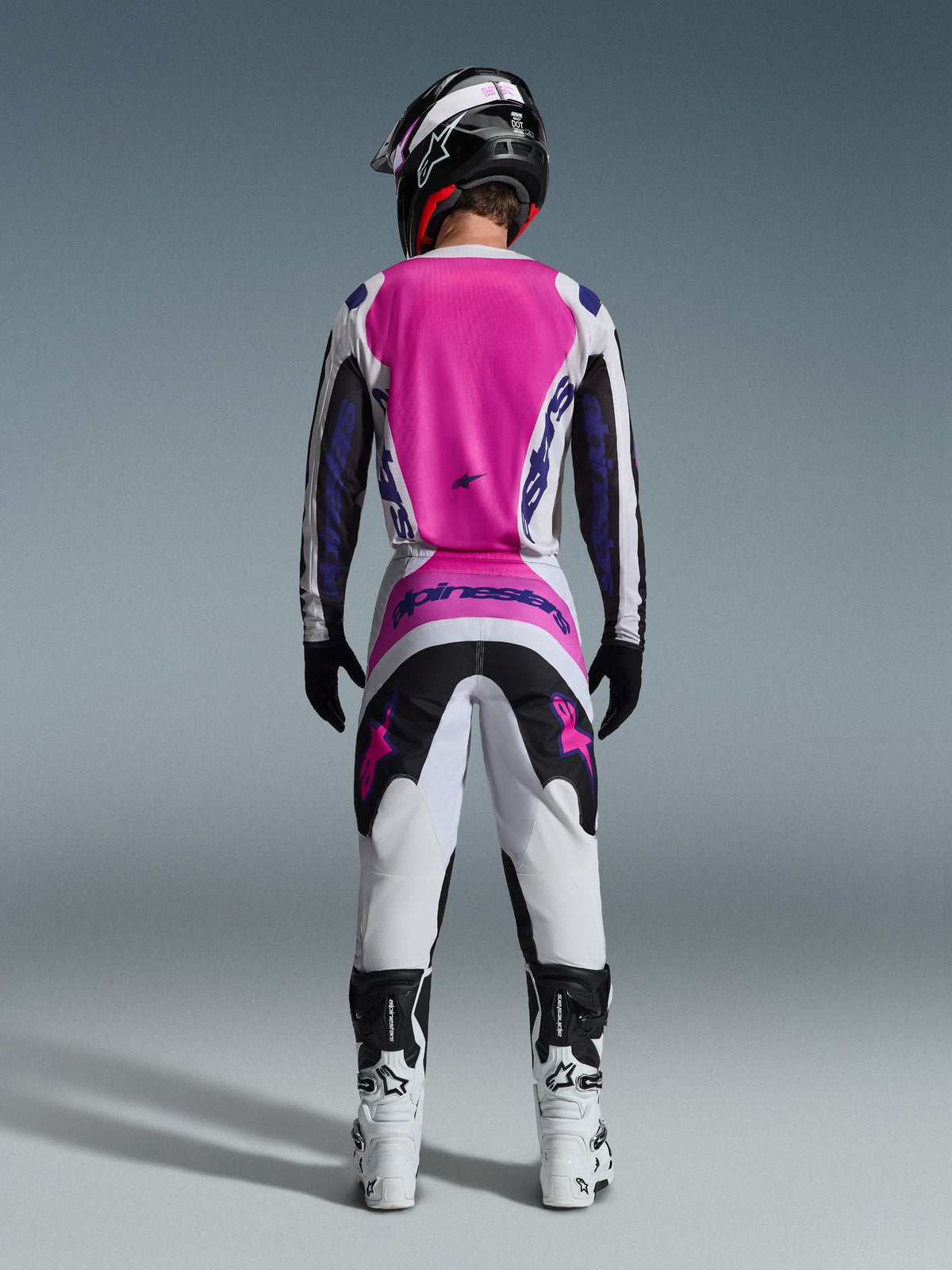 Motocross rider from behind wearing the Alpinestars 2026 Fluid Grid Gear Set, featuring a bright pink and white jersey with black sleeves, matching white pants with black and pink panels, purple Alpinestars branding, white and black racing boots, and a coordinated helmet, designed for off-road performance.