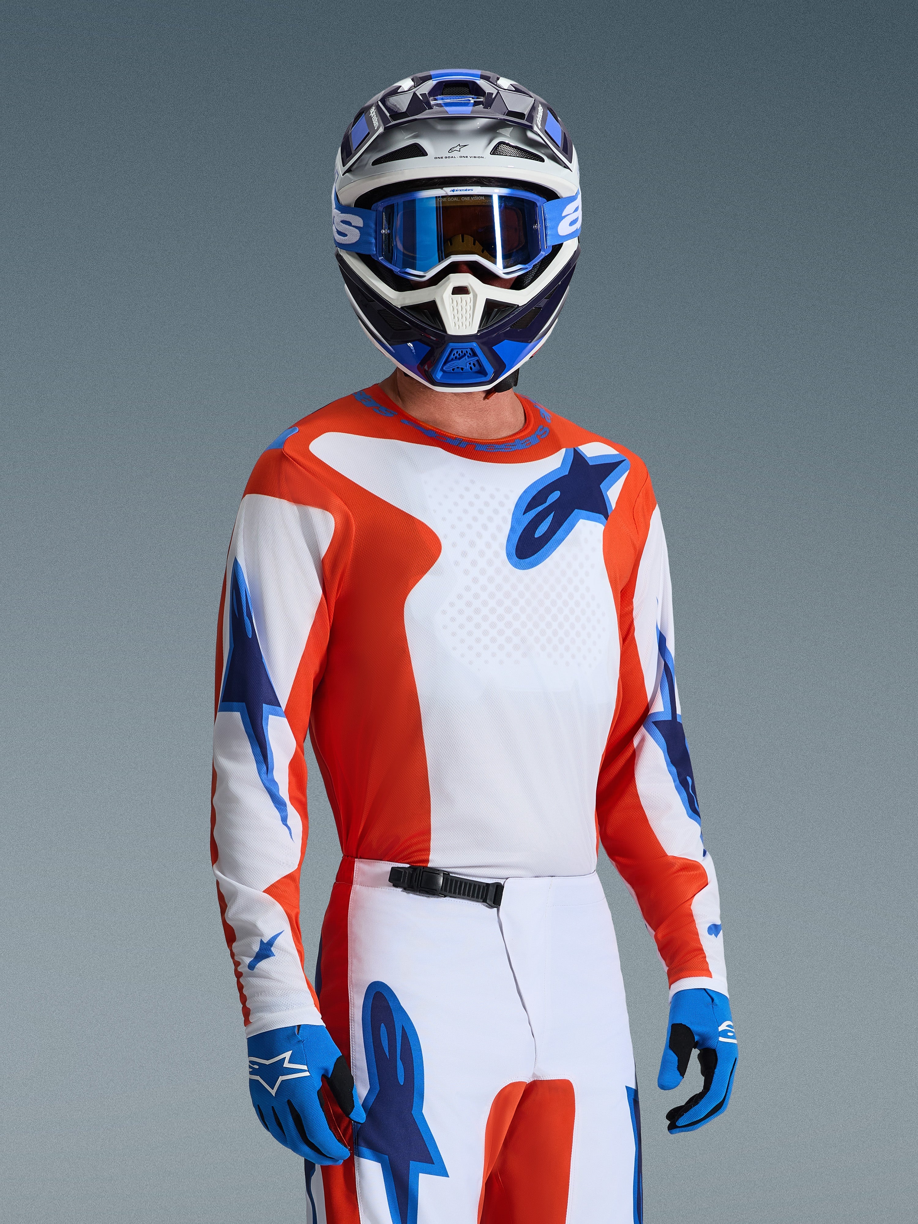 Motocross rider wearing the Alpinestars 2026 Fluid Grid Gear Set, orange and white jersey with blue star logos, white and orange pants with blue accents, blue gloves, blue and white helmet with blue goggles, coordinated athletic racing gear for off-road performance