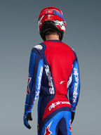 Rückansicht eines Motocross-Fahrers im Alpinestars 2026 Fluid Grid Trikot in Blau und Rot, mit markantem weißem Branding auf den Ärmeln und dem Rücken, kombiniert mit der passenden Motocross-Hose und einem roten Helm für einen kompletten Offroad-Racing-Look