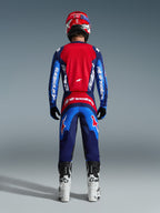 Rückansicht eines Motocross-Fahrers im 2026 Fluid Grid Jersey in Blau und Rot, mit einem großen roten Rückenteil und blauen Ärmeln mit weißem Alpinestars-Schriftzug, kombiniert mit der passenden Hose, weiß-schwarzen Stiefeln und einem roten Helm für ein komplettes Offroad-Rennoutfit.