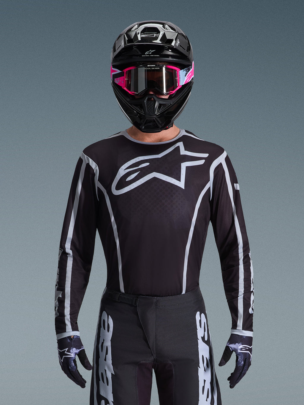 Alpinestars 2026 Fluid Apex Ausrüstungsset, Motocross-Trikot und -Hose in Schwarz und Weiß, mit großem Brustlogo und geometrischen Linien-Designs, getragen mit einem schwarzen Helm und einer pinkfarbenen Brille, Performance-Ausrüstung für Offroad-Motocross-Rennen.