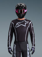 Alpinestars 2026 Fluid Apex Ausrüstungsset, Motocross-Trikot und -Hose in Schwarz und Weiß, mit großem Brustlogo und geometrischen Linien-Designs, getragen mit einem schwarzen Helm und einer pinkfarbenen Brille, Performance-Ausrüstung für Offroad-Motocross-Rennen.