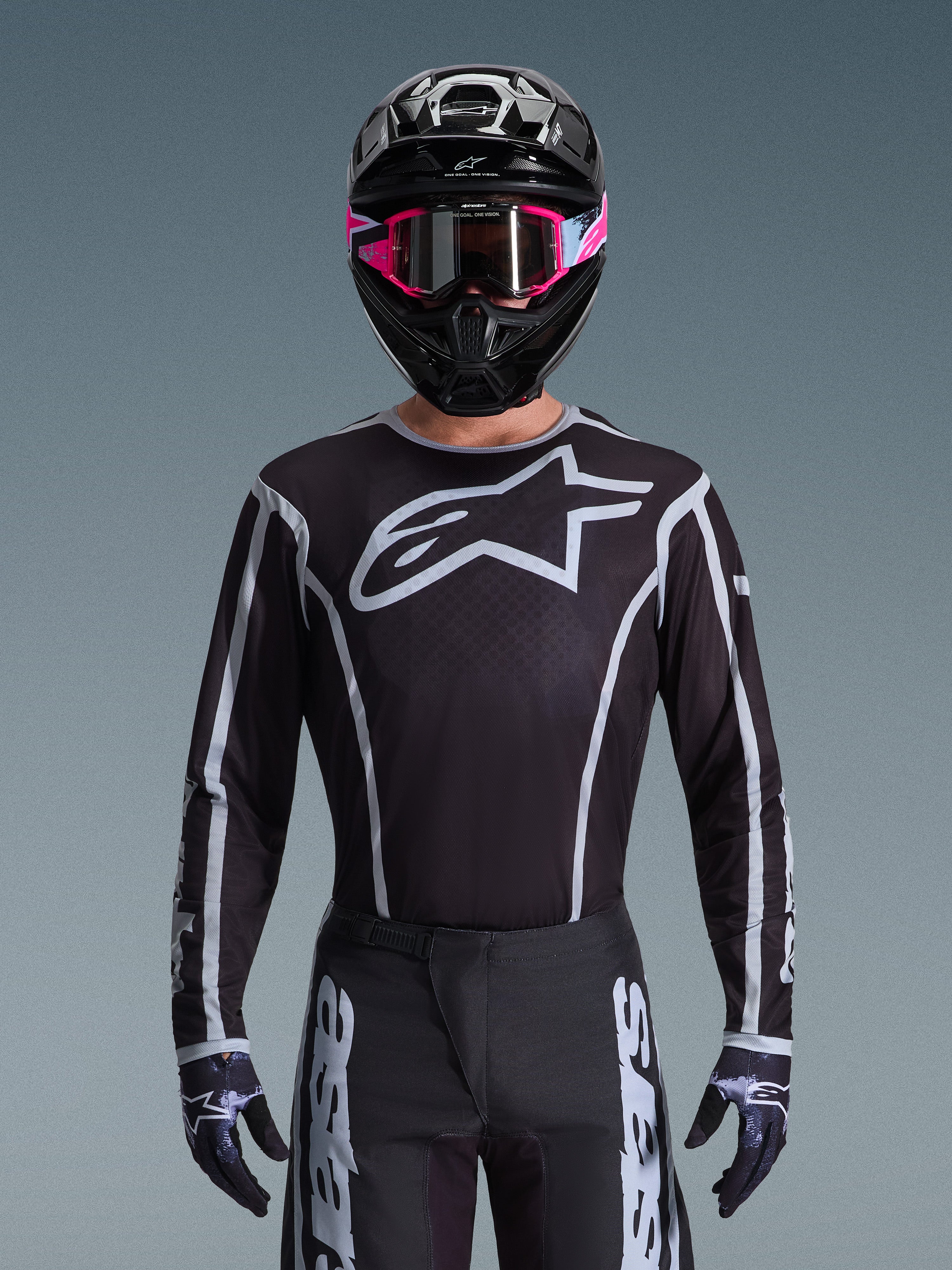 Alpinestars 2026 Fluid Apex Ausrüstungsset, Motocross-Trikot und -Hose in Schwarz und Weiß, mit großem Brustlogo und geometrischen Linien-Designs, getragen mit einem schwarzen Helm und einer pinkfarbenen Brille, Performance-Ausrüstung für Offroad-Motocross-Rennen.