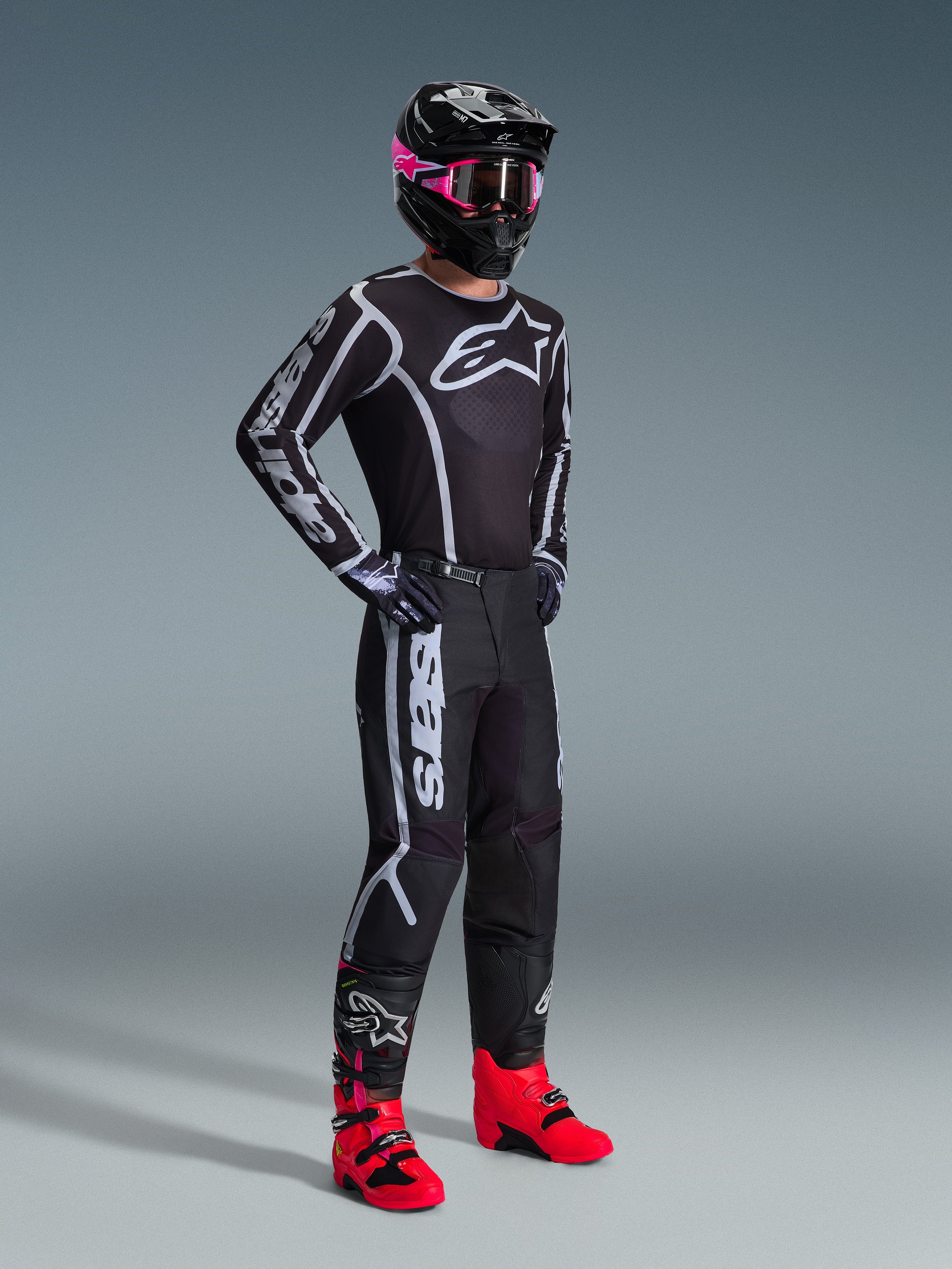 Motocross rider wearing 2026 Fluid Apex Jersey, Black and Gray, paired with matching pants, black helmet with pink goggles, and bright red boots, featuring bold white Alpinestars branding on the chest and sleeves, coordinated professional off-road racing outfit.