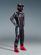 Alpinestars 2026 Fluid Apex Bekleidungsset, Motocross-Fahrer in schwarzem Jersey und Hose mit weiß-grauen Grafikakzenten, schwarzer Helm mit pinker Brille und leuchtend pinken Rennstiefeln, koordiniertes professionelles Motocross-Outfit