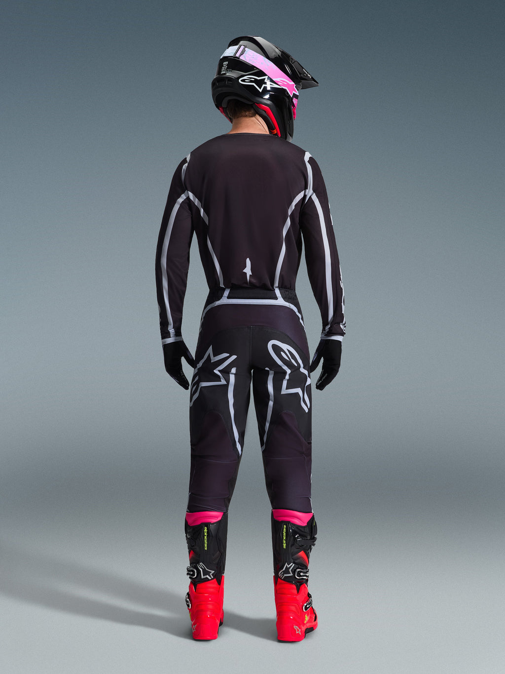 Alpinestars 2026 Fluid Apex Gear Set, Motocross-Fahrer von hinten gesehen, schwarzes Jersey und Hose mit grauen geometrischen Linien und Stern-Logos, schwarzer Helm mit pinkfarbenen Akzenten, leuchtend rote und schwarze Stiefel, abgestimmte Motocross-Rennausrüstung