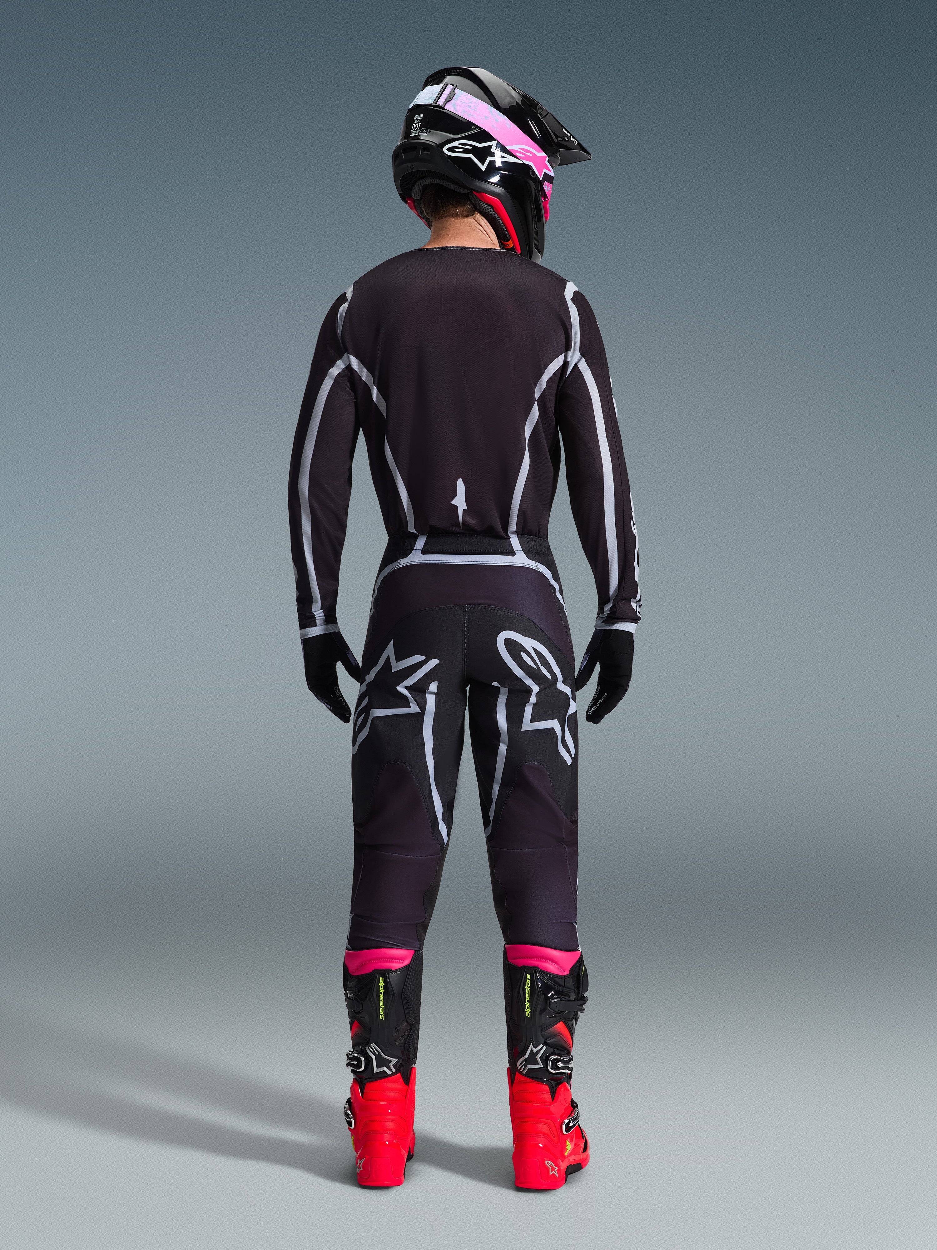 Alpinestars 2026 Fluid Apex Gear Set, Motocross-Fahrer von hinten gesehen, schwarzes Jersey und Hose mit grauen geometrischen Linien und Stern-Logos, schwarzer Helm mit pinkfarbenen Akzenten, leuchtend rote und schwarze Stiefel, abgestimmte Motocross-Rennausrüstung