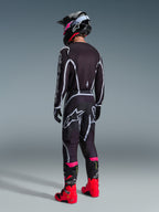 Motocross-Fahrer im Alpinestars 2026 Fluid Apex Gear Set, schwarzes Jersey und Hose mit weißen und grauen geometrischen Linien, Rückansicht, kombiniert mit einem schwarzen Helm mit pinkfarbenen Akzenten und leuchtend roten und pinkfarbenen Rennstiefeln, Studioaufnahme auf grauem Hintergrund.