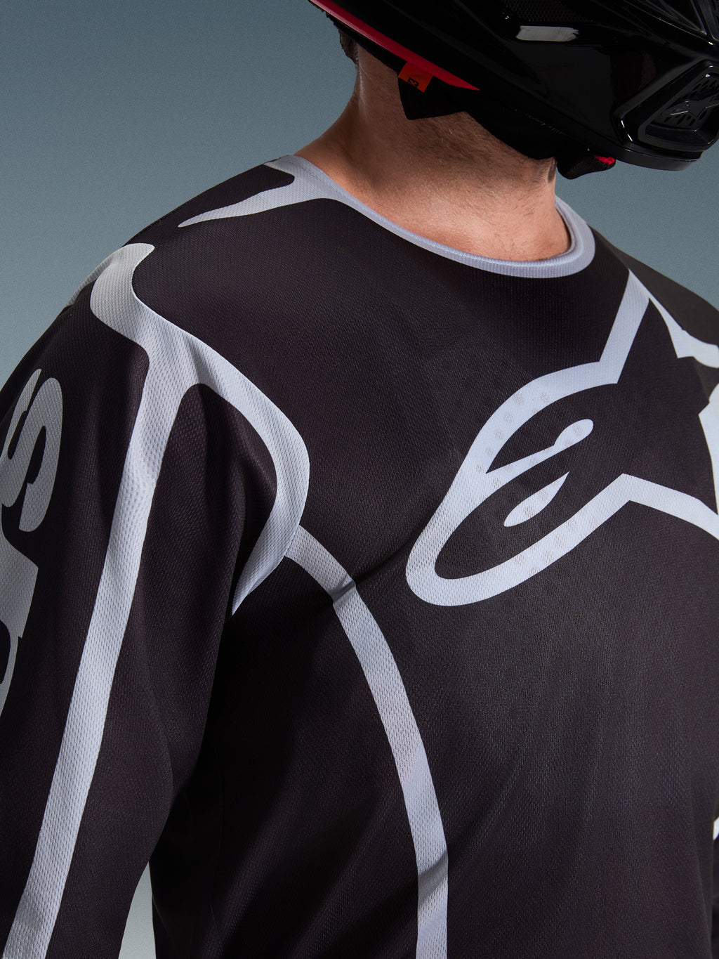 Alpinestars 2026 Fluid Apex Jersey, Motocross-Jersey, Schwarz und Grau, mit großem Alpinestars-Logo auf der Brust, Sportkragen, getragen mit einem schwarzen Helm, Performance-Ausrüstung für den Offroad-Sport