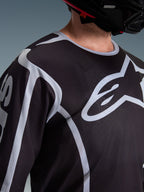 Alpinestars 2026 Fluid Apex Jersey, Motocross-Jersey, Schwarz und Grau, mit großem Alpinestars-Logo auf der Brust, Sportkragen, getragen mit einem schwarzen Helm, Performance-Ausrüstung für den Offroad-Sport