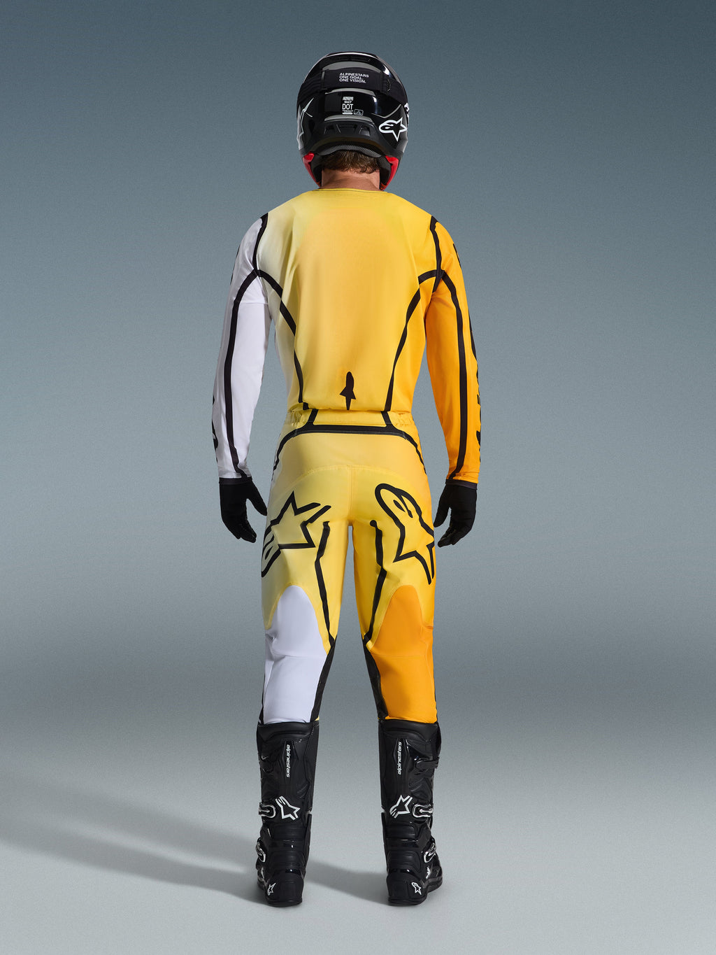 Motocross rider from behind wearing the Alpinestars 2026 Fluid Apex Gear Set, yellow jersey with a white sleeve and black accents, matching yellow pants featuring large black Alpinestars logos on the rear, black helmet, black gloves, and black boots, coordinated outfit for off-road racing