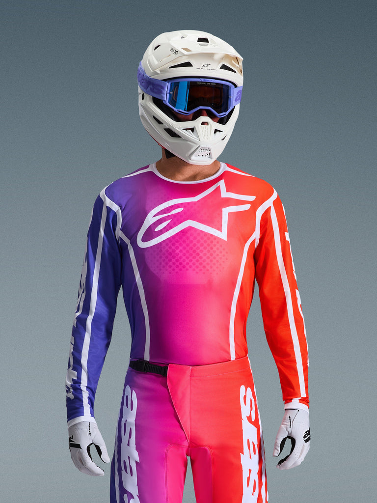 Motocross-Fahrer im 2026 Fluid Apex Trikot, lebhaftes Design mit Farbverlauf in Lila, Pink und Orange, großes weißes Alpinestars-Logo auf der Brust, kombiniert mit weißem Helm, lila Goggles und weißen Handschuhen für einen kompletten Offroad-Rennlook.