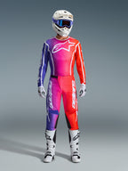 Motocross-Fahrer in kompletter Alpinestars 2026 Fluid Apex Rennausrüstung, mit Trikot und Hose in einem lila, pinken und orangefarbenen Farbverlauf. Das abgestimmte Outfit umfasst ein weißes Alpinestars-Logo auf der Brust, einen weißen Helm mit blauer Brille und weiße Stiefel, entwickelt für Offroad-Performance.