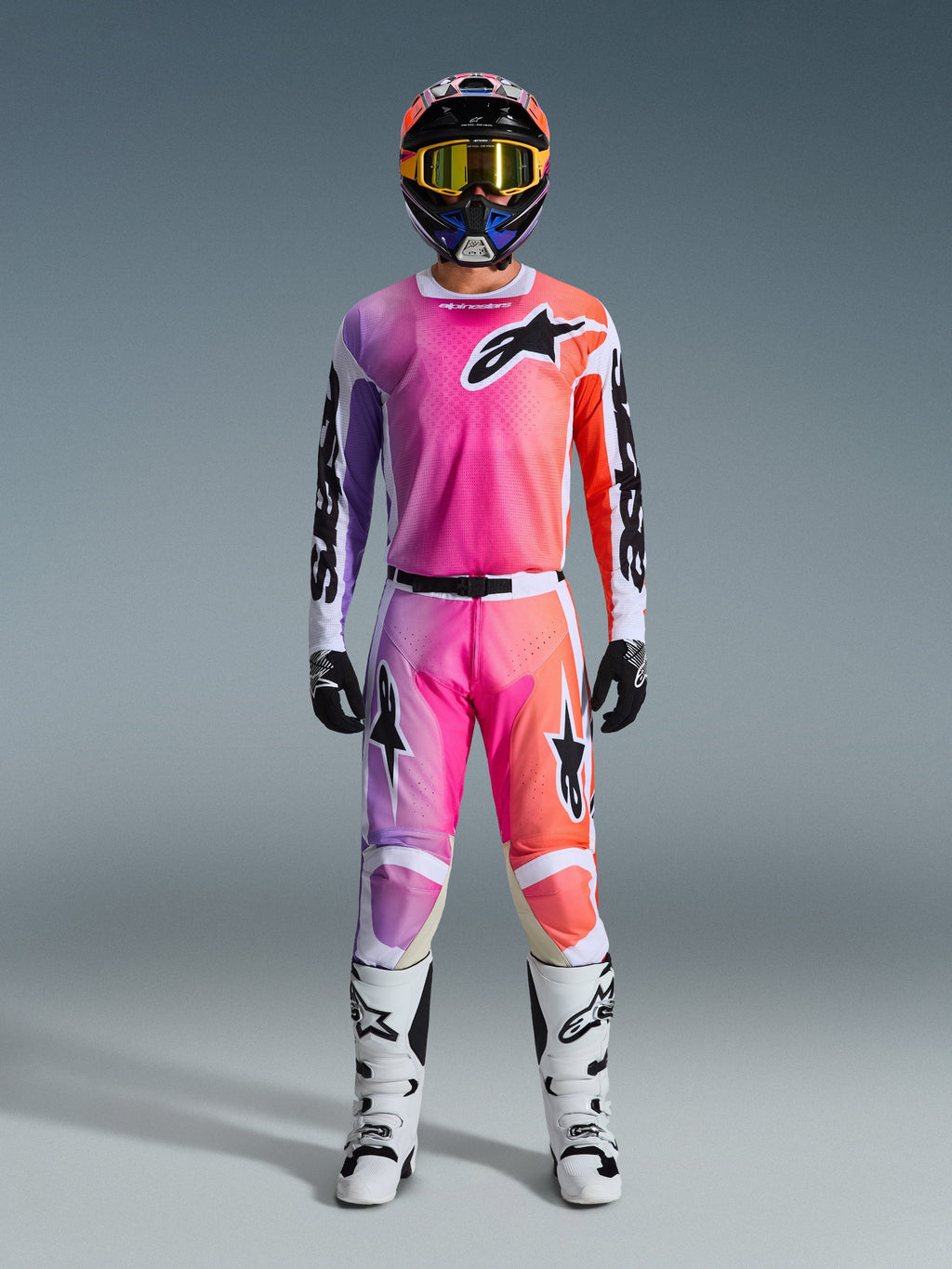 Motocross-Fahrer in kompletter Alpinestars 2026 Racer Air Portl Rennausrüstung, mit einem pinken, lila und orangefarbenen Farbverlauf auf Hose und Trikot, kombiniert mit einem passenden Helm, gelber Schutzbrille, weißen Stiefeln und schwarzen Handschuhen für einen abgestimmten Offroad-Look.