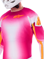 Alpinestars Limited Edition Supertech AFD MIG Jersey, Motocross-Jersey mit Farbverlauf in Pink Fluo und Weiß, mit orangefarbenen Astars-Logos und Ärmelakzenten, getragen von einem Fahrer mit weißem Helm, athletische Racing-Passform.