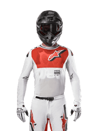 Motocross-Fahrer im Alpinestars 2024 Supertech Ward Trikot in White Hot Orange, weißes Stretch-Mesh-Trikot mit markanter roter Brustgrafik und Logo, schwarzer Helm mit dunkler Brille, weiße Handschuhe, professionelle Ausrüstung für Offroad-Performance