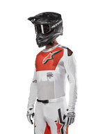 Alpinestars 2024 Supertech Ward Trikot, Motocross-Trikot, Weiß und Hot Orange, mit rotem Brustpanel und weißem Stern-Logo, Langarm mit lasergeschnittenen Perforationen, getragen mit einem schwarzen Helm und weißen Handschuhen, athletische Rennpassform Motocross-Ausrüstung