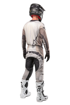 Motocross rider from the back wearing the 2024 Techstar Pneuma Jersey in Dark Sand Iron Dust Gray, matching motocross pants with bold branding, black helmet, and white racing boots, coordinated professional off-road gear.