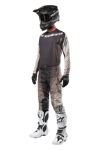 Motocross rider wearing complete racing gear, 2024 Techstar Pneuma Jersey in Dark Sand and Iron Dust Gray, matching patterned motocross pants, black helmet with goggles, white and black boots, black gloves, coordinated outfit for off-road racing