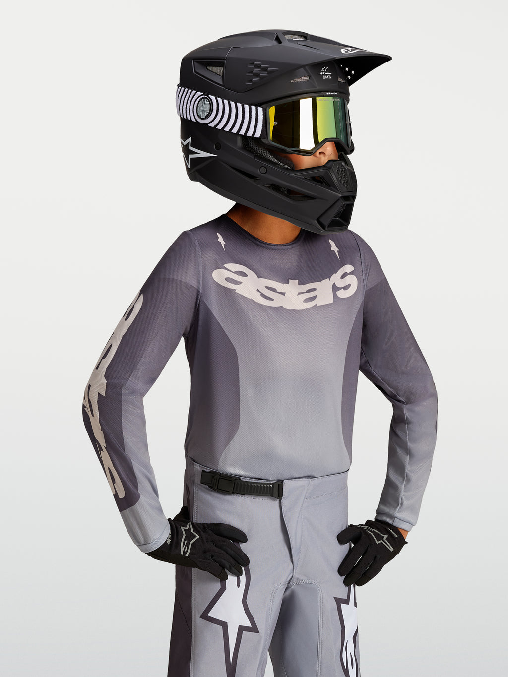Alpinestars Fluid Jugend-Trikot Amber 2027, Jugend-Motocross-Trikot, Grau und Sand, leichtes atmungsaktives Material, getragen mit schwarzem Rennhelm und Handschuhen.