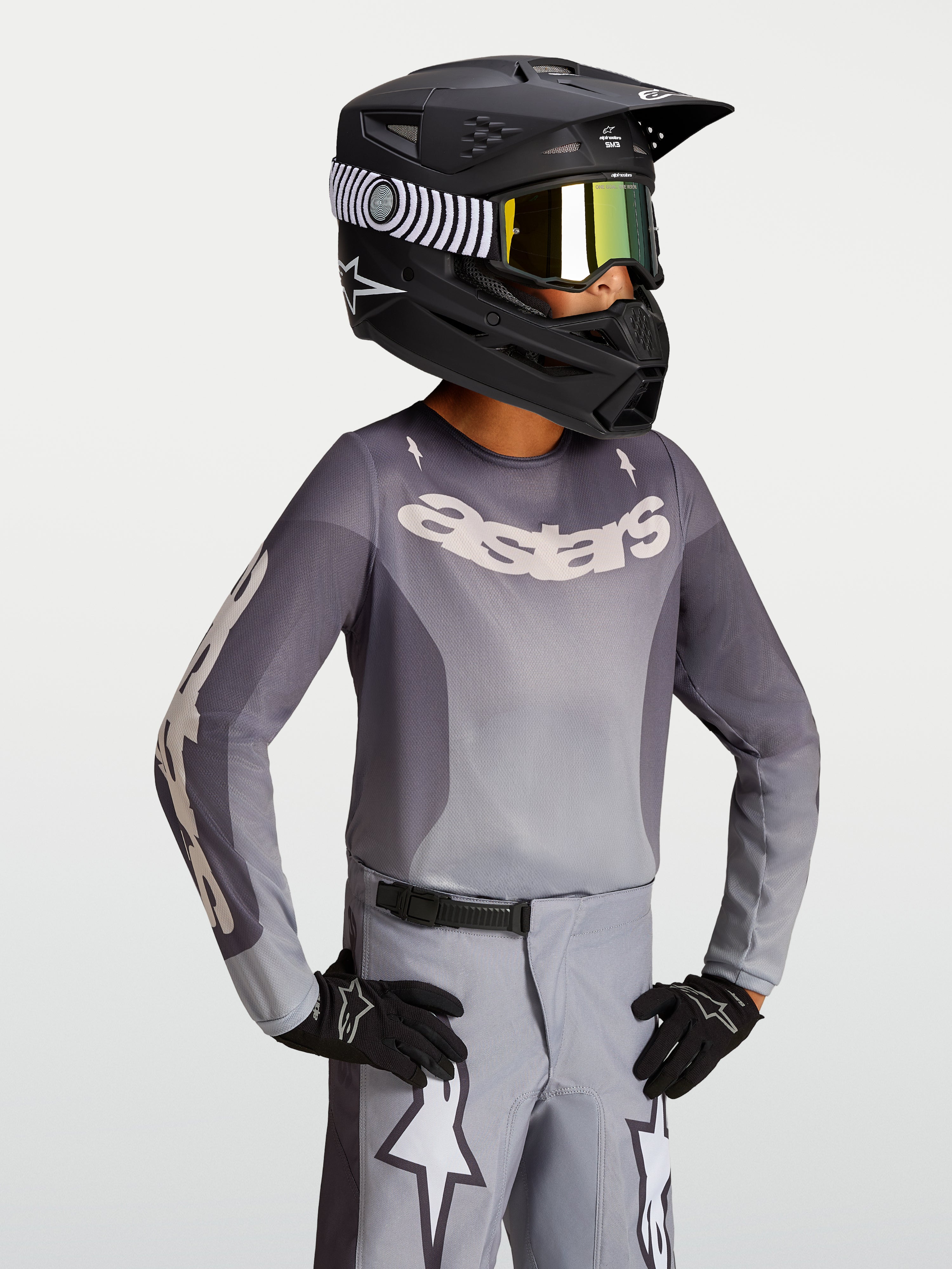 Alpinestars Fluid Jugend-Trikot Amber 2027, Jugend-Motocross-Trikot, Grau und Sand, leichtes atmungsaktives Material, getragen mit schwarzem Rennhelm und Handschuhen.