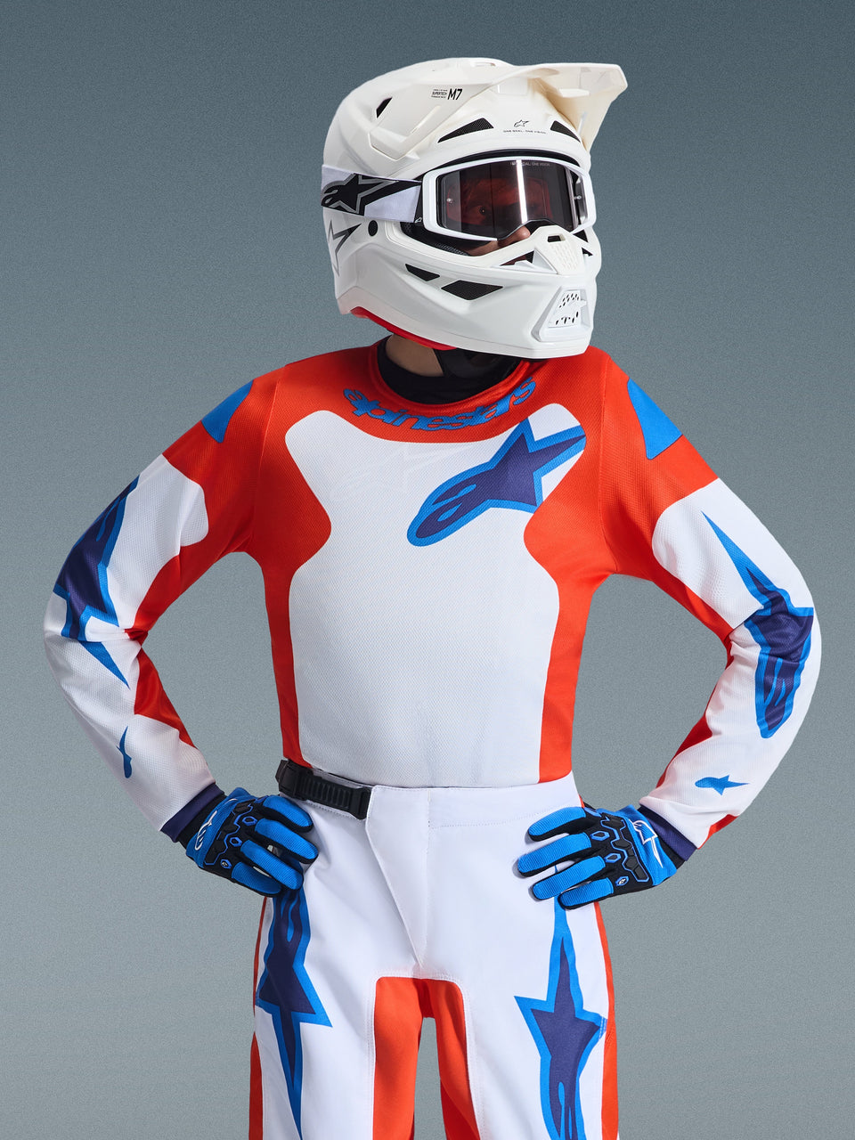 Youth motocross rider wearing the Alpinestars 2026 Youth Fluid Grid Jersey, Orange and UCLA Blue with a white center panel and blue logo, paired with matching pants, blue and black gloves, and a white helmet with goggles, complete racing gear set for off-road riding.